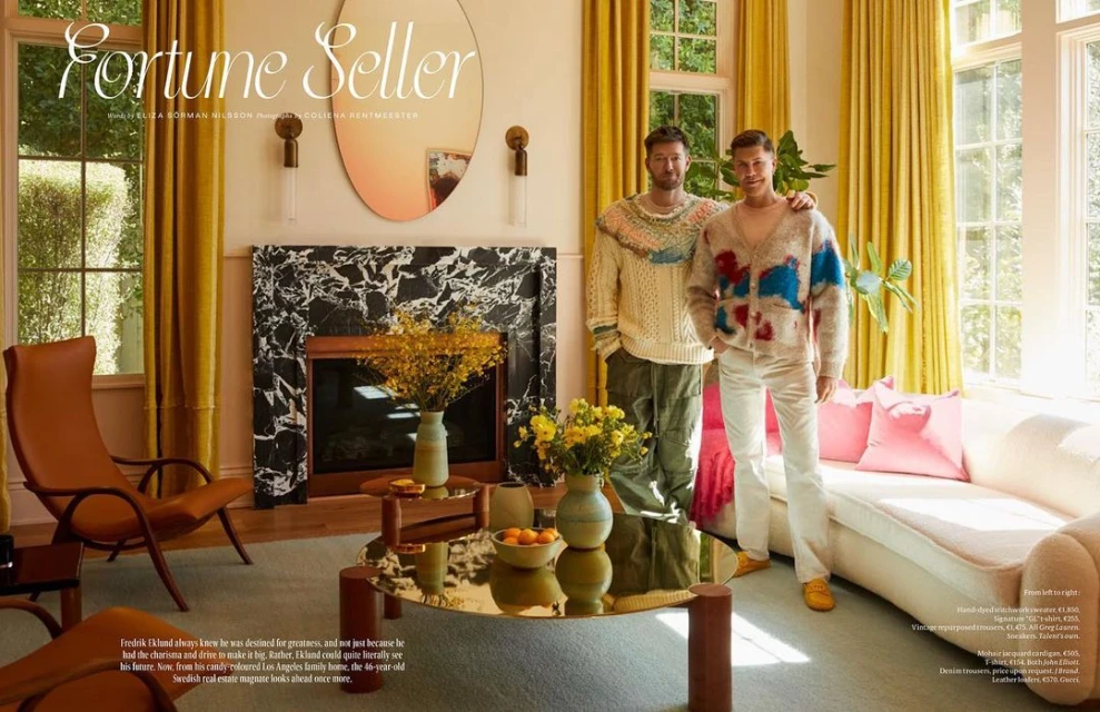 Two adult men stand in an elegant living room with yellow curtains, a marble fireplace, a round glass table, and a white sofa with pink pillows.