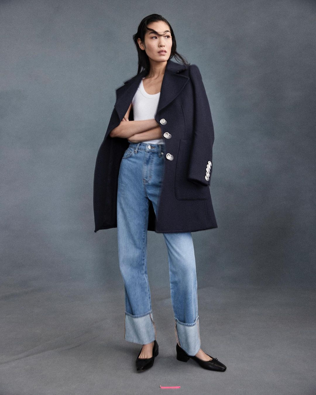 A full-length shot captures an Asian woman posing in a navy coat, white top, and blue jeans against a gray backdrop in a studio setting.