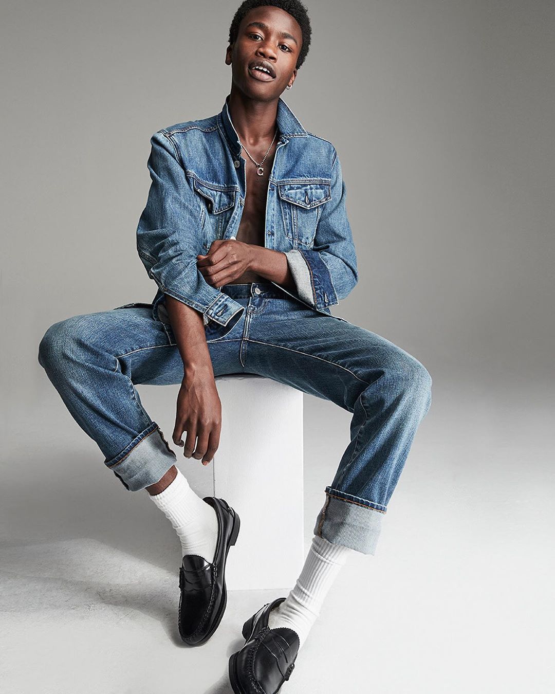 A young African-American man sits on a white cube in a photo studio, posing in a denim jacket, jeans, white socks, and black loafers.