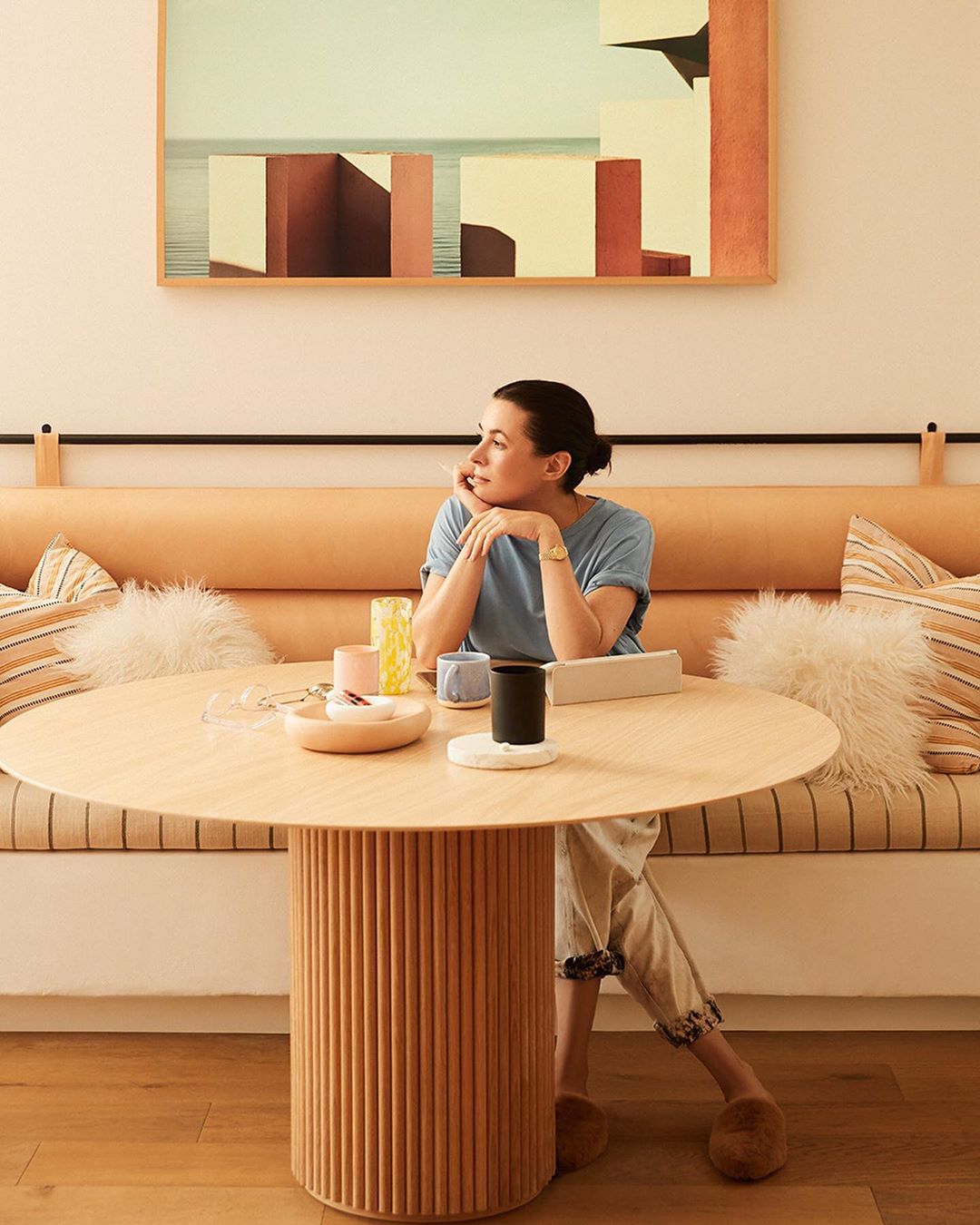 Woman sits at round table with neutral decor. An abstract painting hangs on the wall behind her. She's wearing a blue shirt and tan pants.