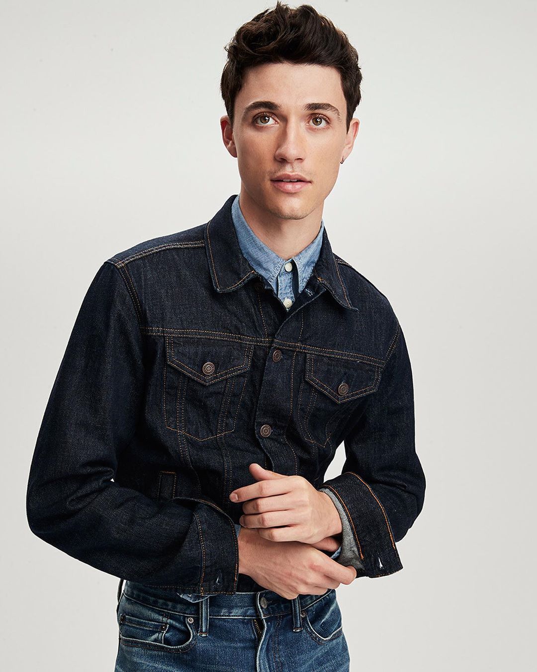 A young adult man with dark hair poses in a studio wearing a denim jacket and light blue shirt, against a white backdrop in a medium shot.