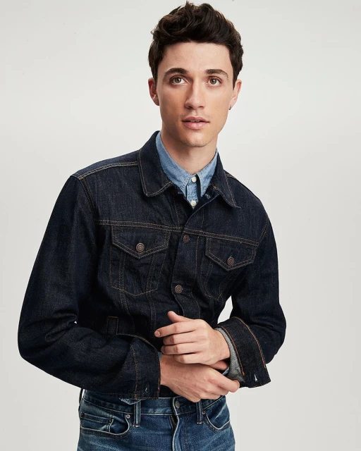 A young adult man with dark hair poses in a studio wearing a denim jacket and light blue shirt, against a white backdrop in a medium shot.
