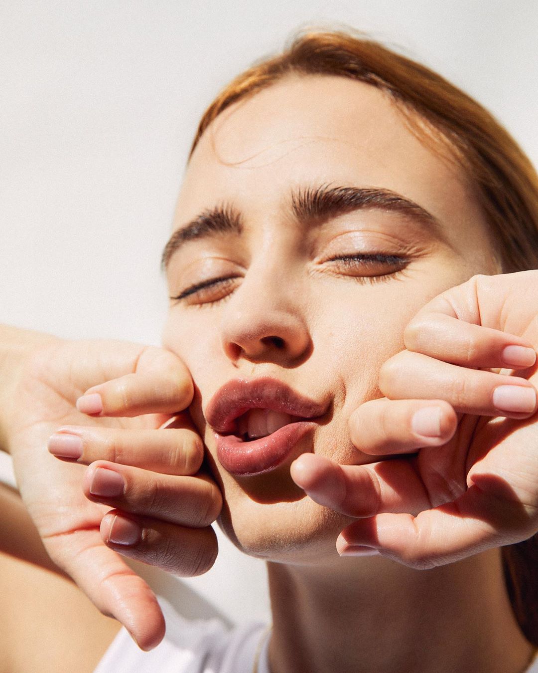 Portrait of an adult woman with closed eyes, pouting lips and hands framing her face against a bright white background, beauty shot.