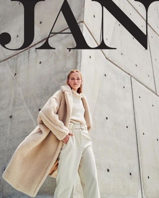 Fashion shot: woman in beige coat poses against a concrete wall with black text overlay. Neutral colors, fashion editorial style.
