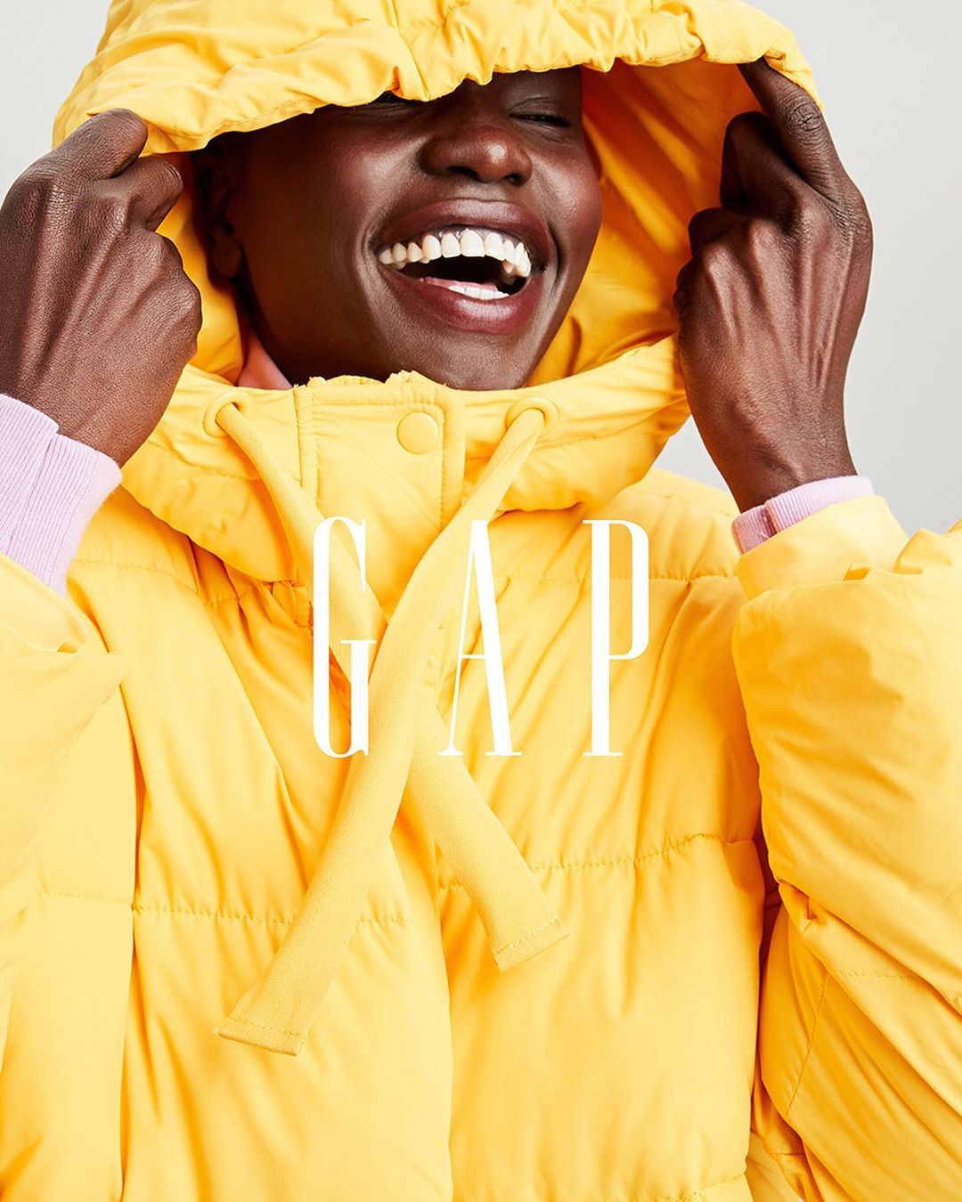 Smiling Black woman wears bright yellow puffer jacket with hood indoors; GAP logo across front. Fashion, beauty, lifestyle concept.