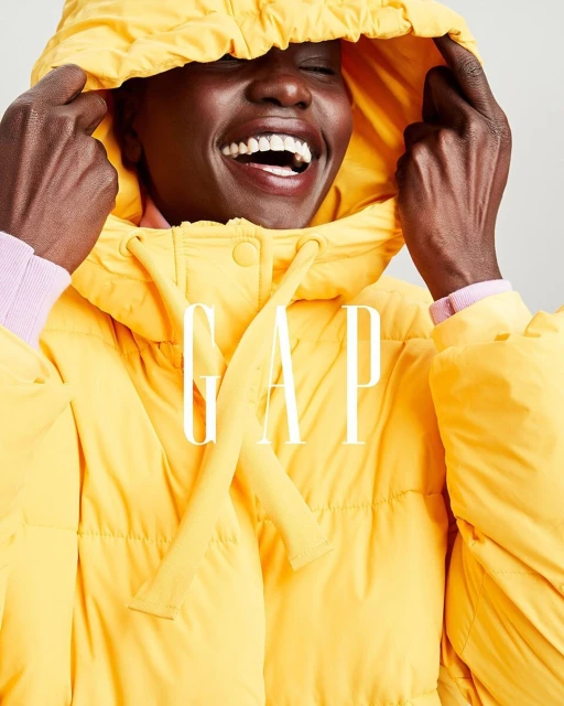 Smiling Black woman wears bright yellow puffer jacket with hood indoors; GAP logo across front. Fashion, beauty, lifestyle concept.