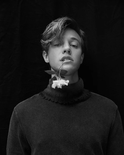 Black and white portrait of a young adult male with flower in mouth, wearing a turtleneck sweater against a dark studio background.