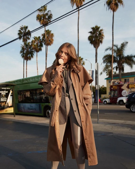 Woman in trench coat stands on a city street eating a lollipop with palm trees, bus, and buildings in background, daytime.