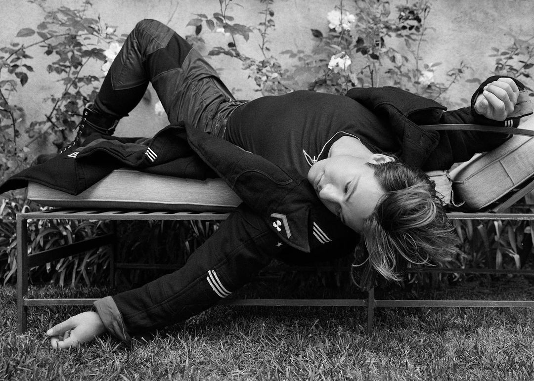 Black and white shot of a person lounging on a metal outdoor bench in a garden, wearing denim and a jacket with grass and plants around.