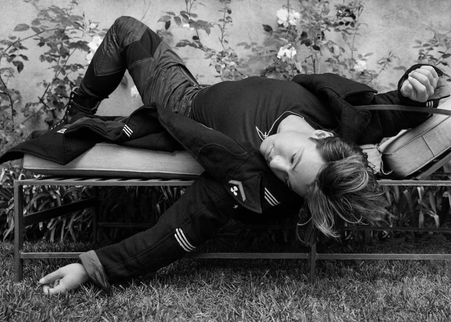 Black and white shot of a person lounging on a metal outdoor bench in a garden, wearing denim and a jacket with grass and plants around.