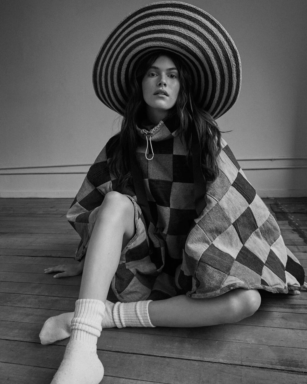 A black and white portrait of a young woman wearing a large, striped hat and checkered dress, sitting on a wooden floor.