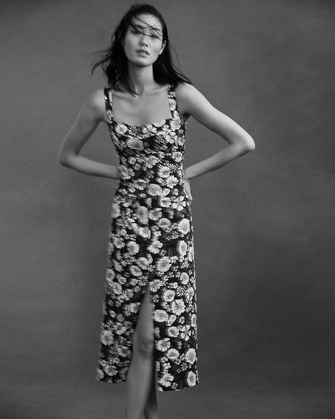 Monochrome portrait of an Asian woman in a floral print dress with a front slit, posing against a neutral grey backdrop in a studio.