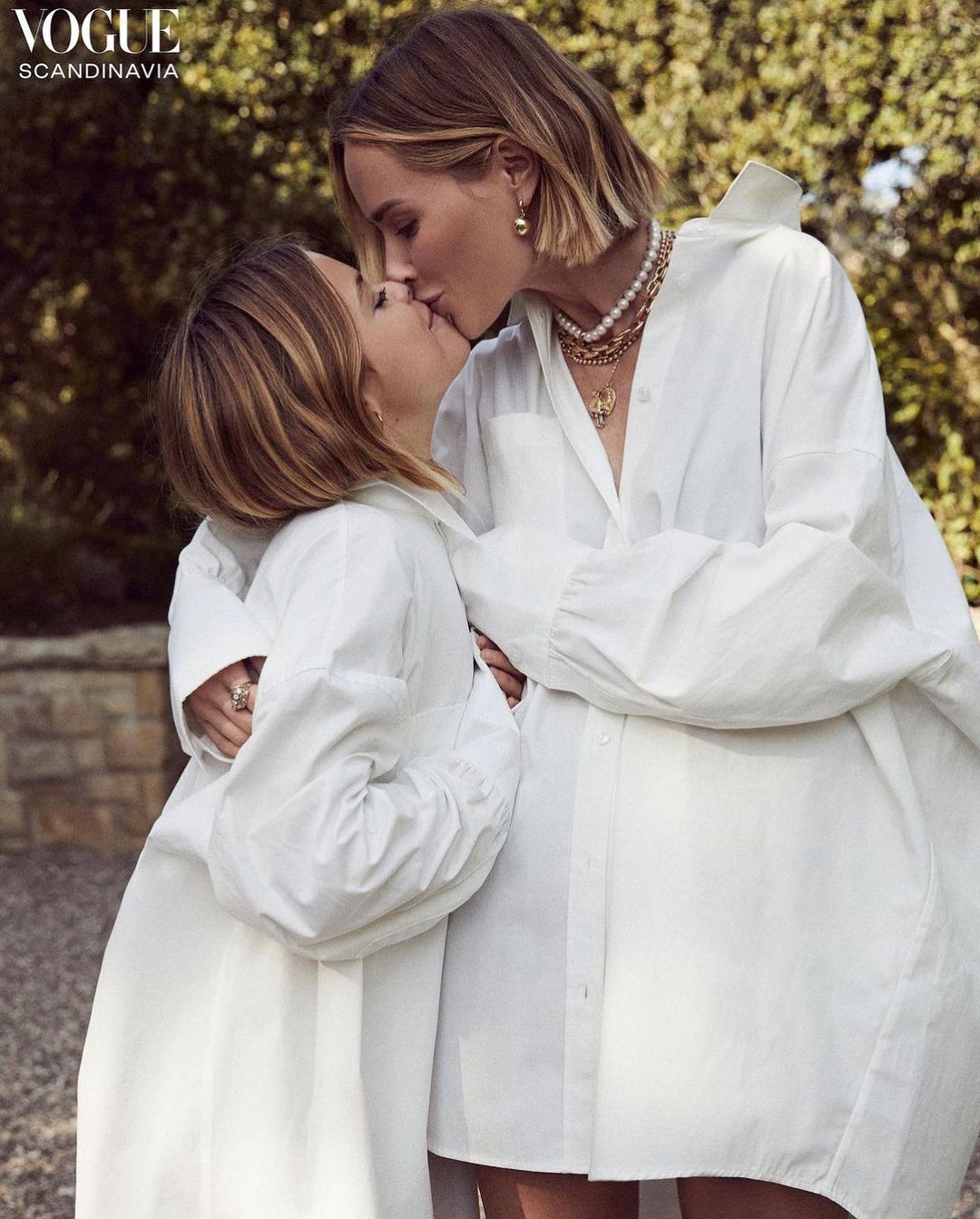 Two women in white oversized shirts embrace and kiss outdoors in a fashion editorial style. Vogue Scandinavia logo visible.