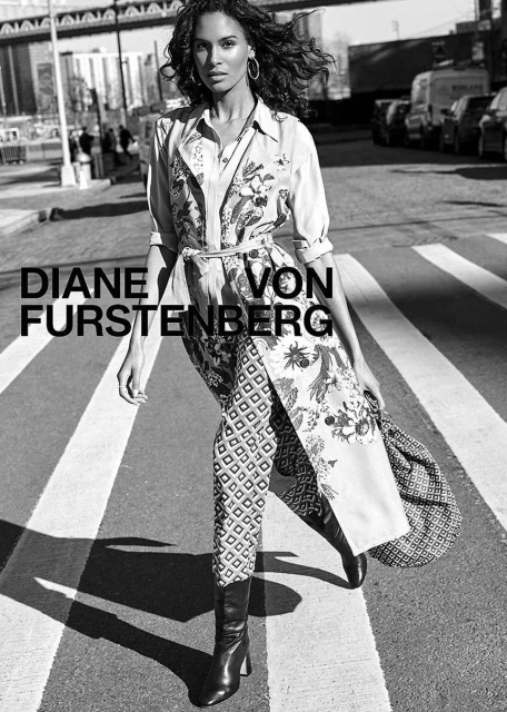 Black and white full shot of a fashionable adult female posing on a city crosswalk wearing patterned clothing and boots. Cars in background.