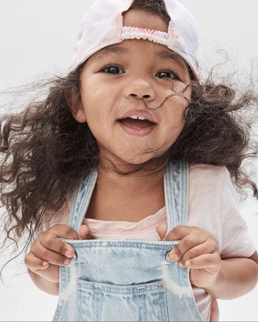 Adorable child with curly hair smiles wearing denim overalls and a baseball cap, set against a clean white background in a close-up shot.