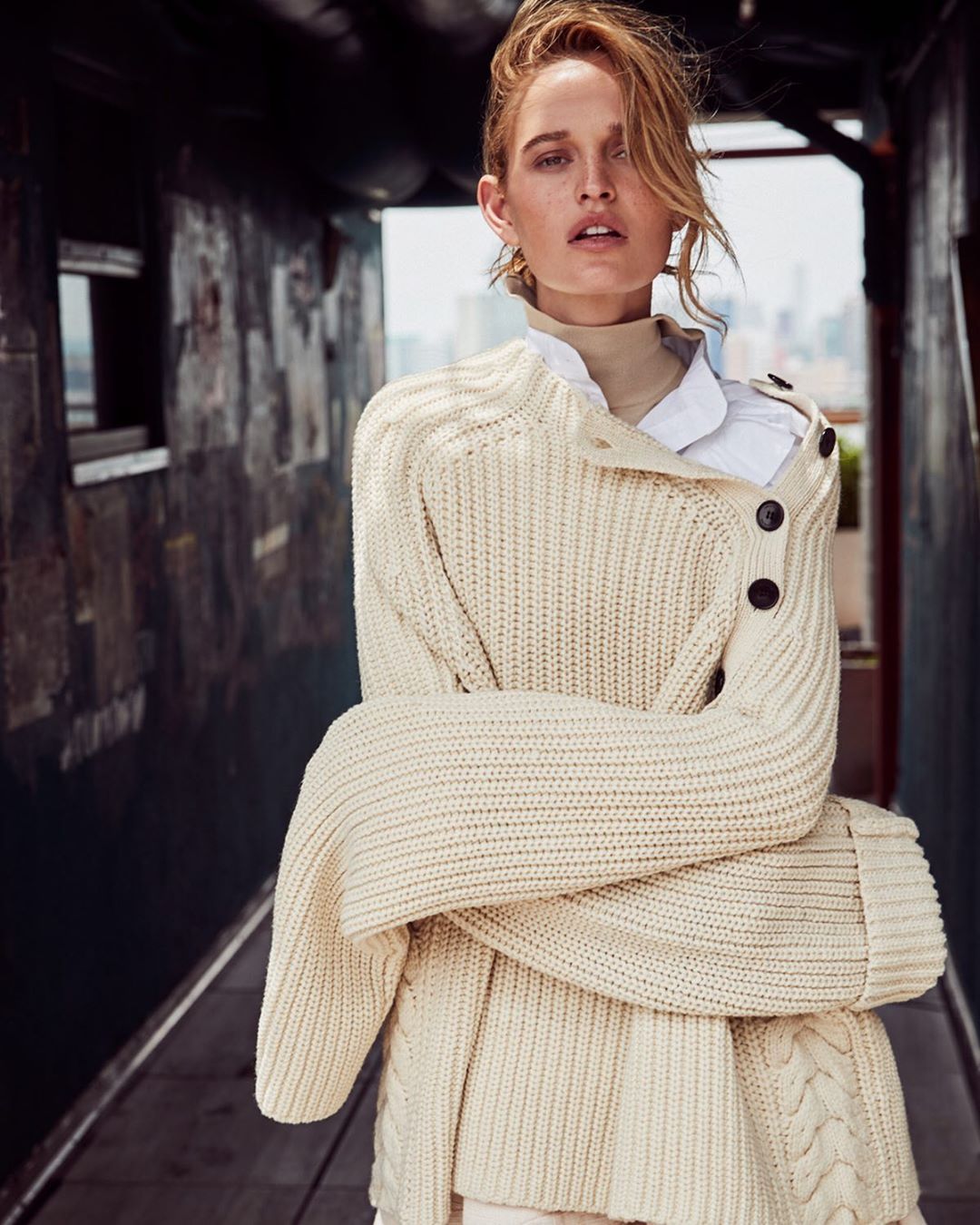 Fashion portrait of a blonde female model in an ivory knit sweater, posing with arms crossed against an urban backdrop on an overcast day.