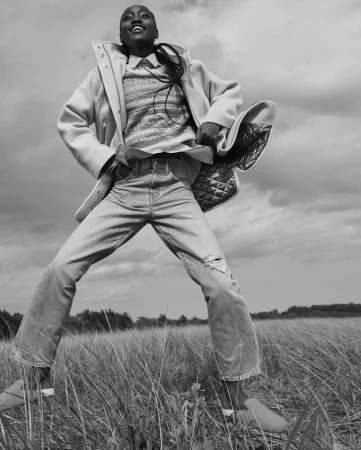 Low-angle black-and-white portrait of a smiling Black model wearing a patterned sweater, open coat, and jeans standing in a tall grass meadow.
