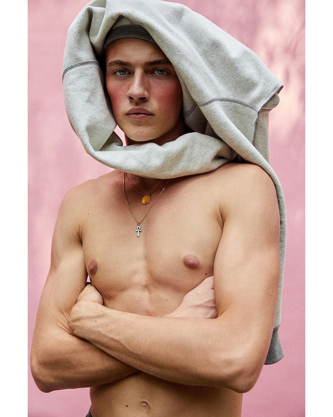 Shirtless young man with arms crossed wearing a grey sweatshirt draped over his head against a pink textured wall.