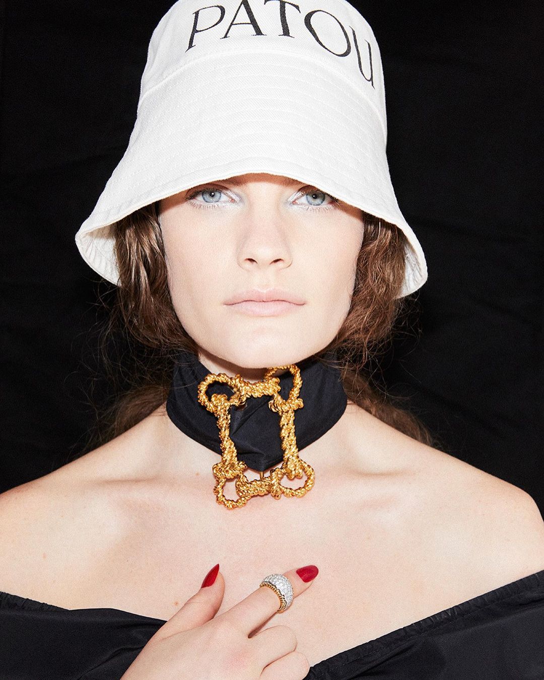 Woman wearing Patou bucket hat and gold choker against a black background. Her manicured hand displays a ring. Beauty shot.