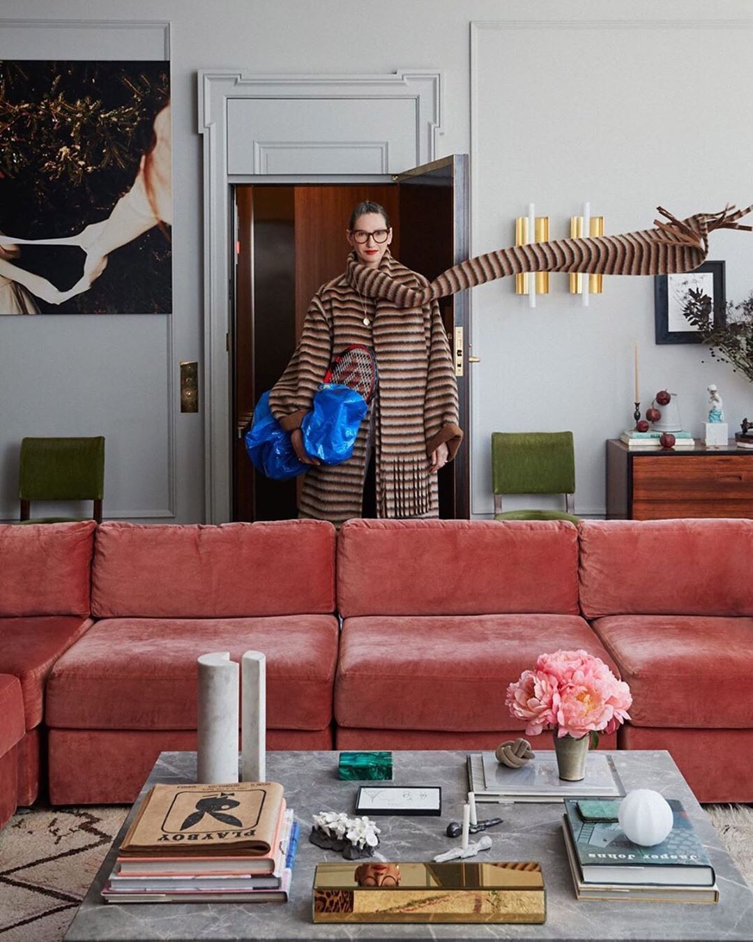 Fashionable adult woman wearing glasses, a striped coat, and holding blue bags, posing in a stylishly decorated living room.