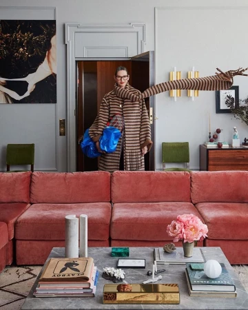 Fashionable adult woman wearing glasses, a striped coat, and holding blue bags, posing in a stylishly decorated living room.