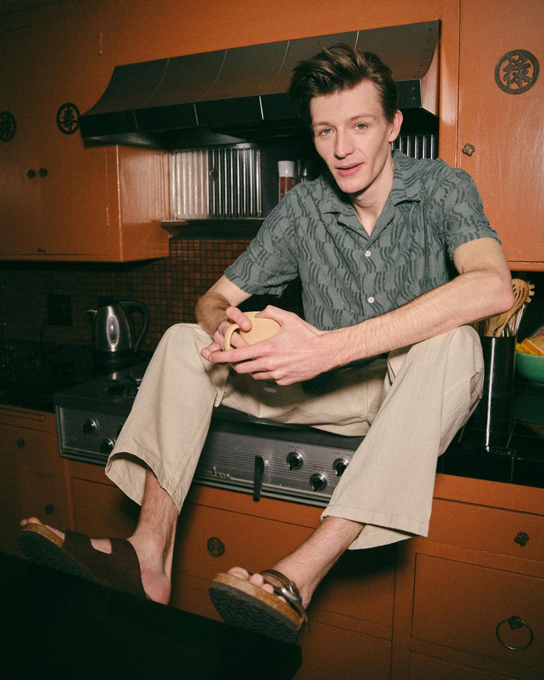 Young man sitting on kitchen counter wearing casual clothing and sandals, holding a mug, with orange cabinets and a kettle in the background.