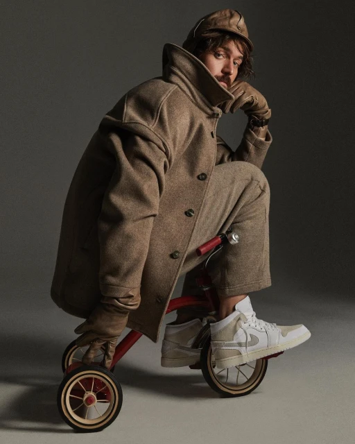Man in tweed coat and hat sits on a child's tricycle in a studio with a gray background, wearing white sneakers and gloves. 