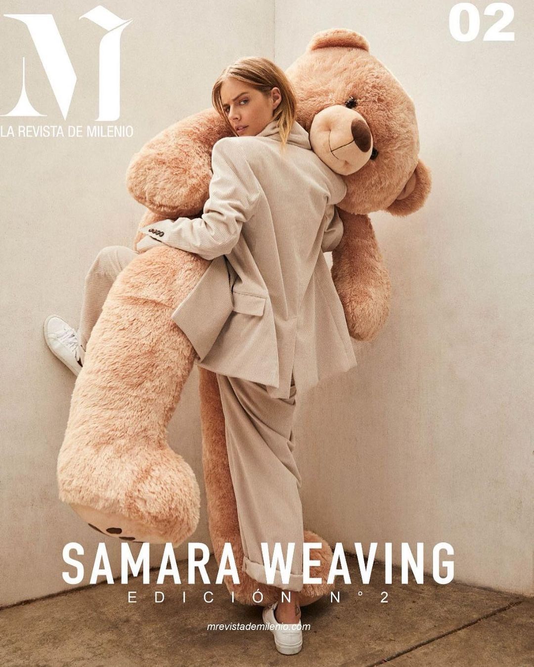 Editorial fashion shot of blonde woman in a cream-colored suit, embracing a large teddy bear, on a magazine cover with white background.