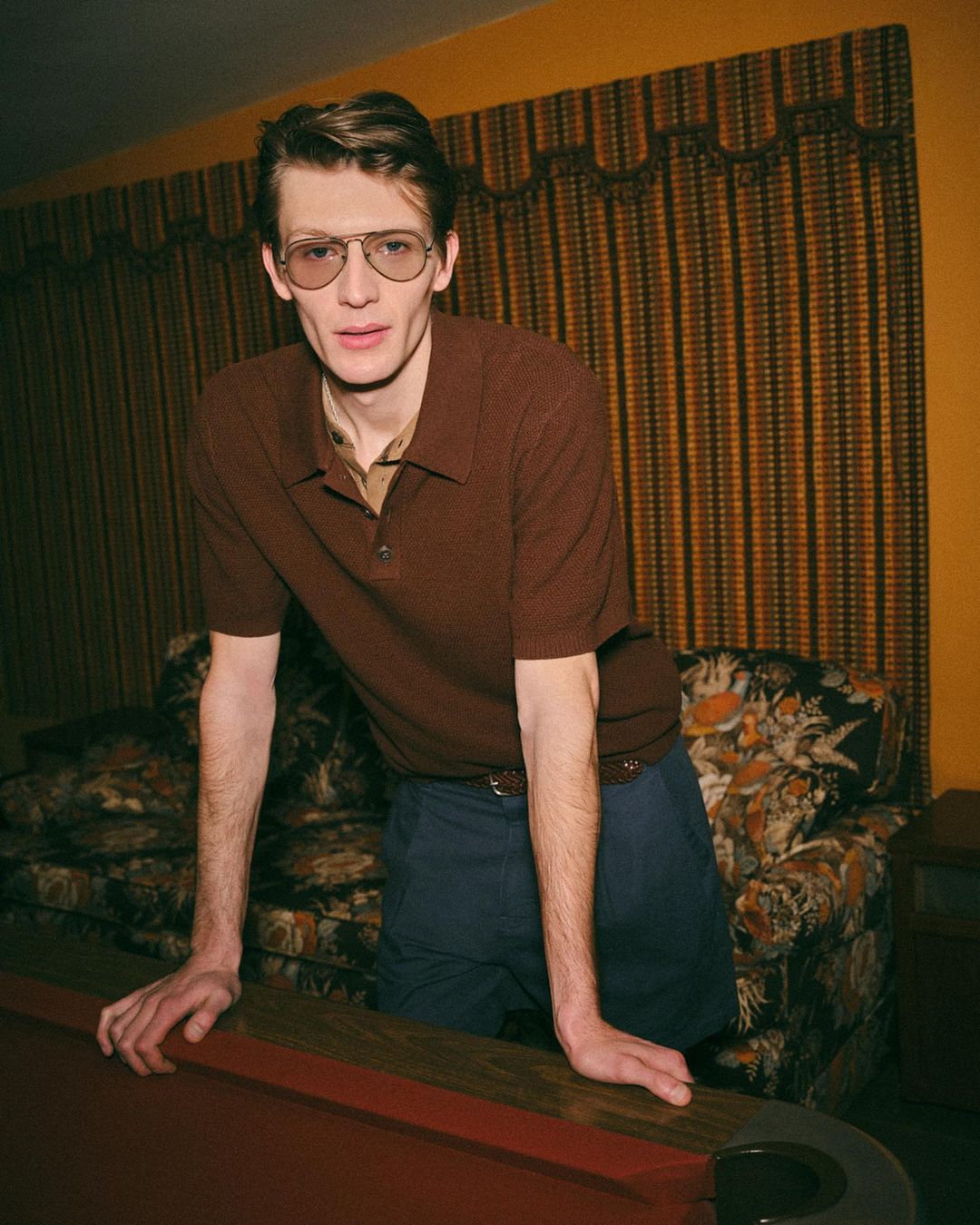An adult male wearing glasses and a brown polo shirt poses indoors in front of a floral couch and a pool table with a neutral-toned wall.