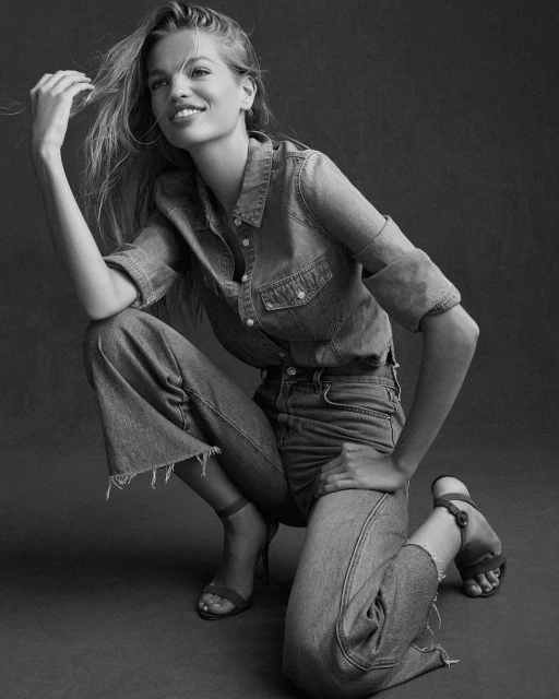Black and white portrait of a Caucasian woman in a denim outfit crouching, smiling, and holding her hair.