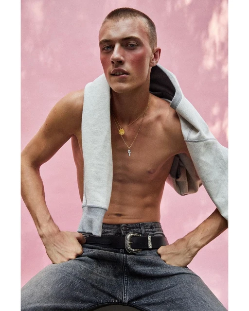 Male fashion model poses shirtless in jeans and a hoodie against a pink background. Gold necklaces accent the look.
