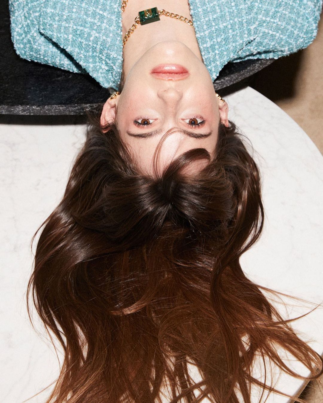 Upside-down portrait of a woman with long brunette hair, wearing a blue tweed blazer and gold jewelry, against a marble table.