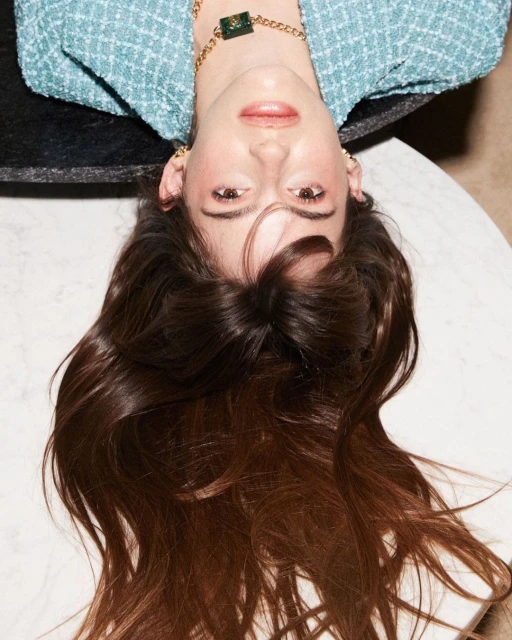 Upside-down portrait of a woman with long brunette hair, wearing a blue tweed blazer and gold jewelry, against a marble table.