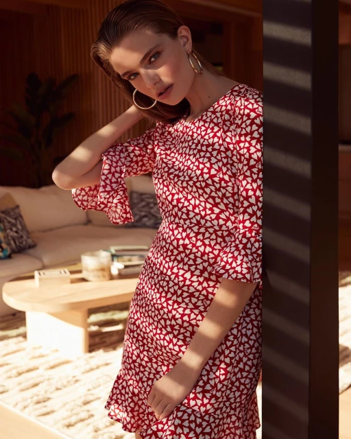 A fashionable young woman in a red and white heart-print dress poses in an interior setting with a neutral color palette and soft lighting.