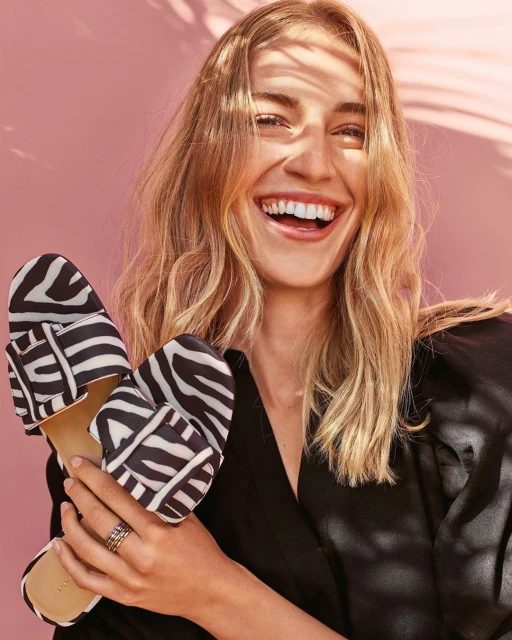 A smiling woman in a black top holds two shoes with a black and white zebra print in front of a pink background with soft shadows.