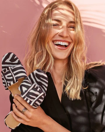 A smiling woman in a black top holds two shoes with a black and white zebra print in front of a pink background with soft shadows.