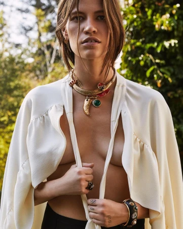 Model wearing a cream cape, layered gold statement necklaces, and dark bracelets outdoors with green foliage.