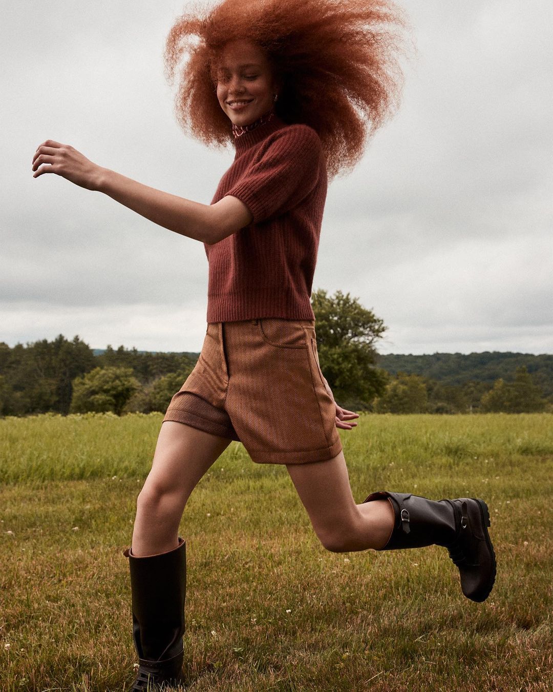 A woman with red hair runs through a grassy field wearing a brown sweater, brown shorts and black boots with cloudy sky overhead.