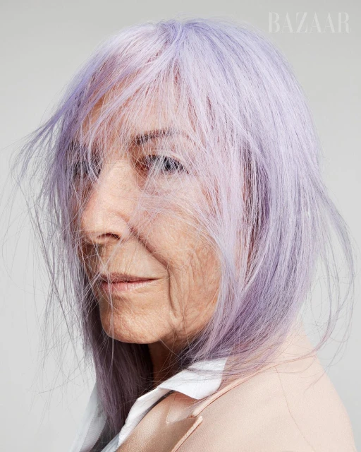 Close-up of a mature woman with lavender-colored hair, wearing a peach blazer and white shirt against a neutral gray background.