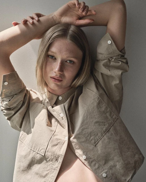 Portrait of a blonde woman with freckles wearing a tan button-down shirt, posing with her arms raised against a light background.