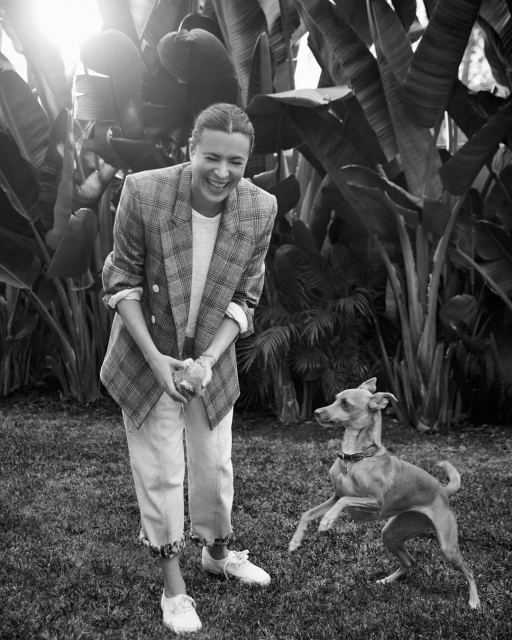 Black and white shot of an adult woman playing with a dog outdoors on a grassy lawn, surrounded by large leafy plants.