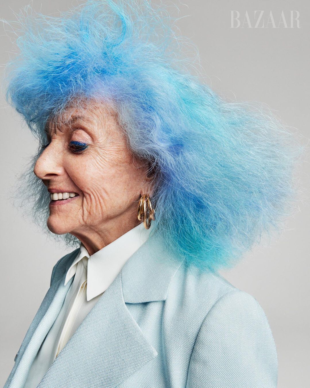 Profile portrait of an elderly woman with brightly dyed blue hair wearing a light blue blazer and large gold hoop earrings, smiling gently.