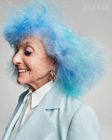 Profile portrait of an elderly woman with brightly dyed blue hair wearing a light blue blazer and large gold hoop earrings, smiling gently.