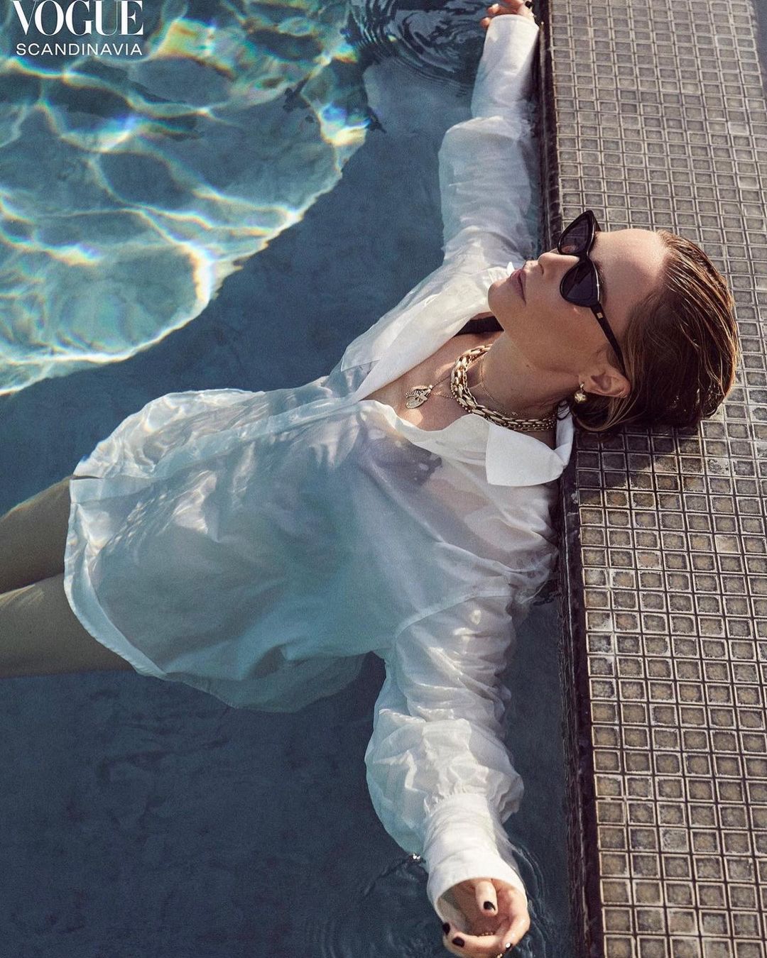 Woman in a white shirt and sunglasses relaxes in a swimming pool with light reflecting on the water, photographed for Vogue Scandinavia.