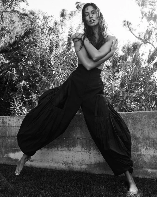 Black-and-white portrait of a young adult woman with long hair posing outdoors in flowing black pants, standing in grass.