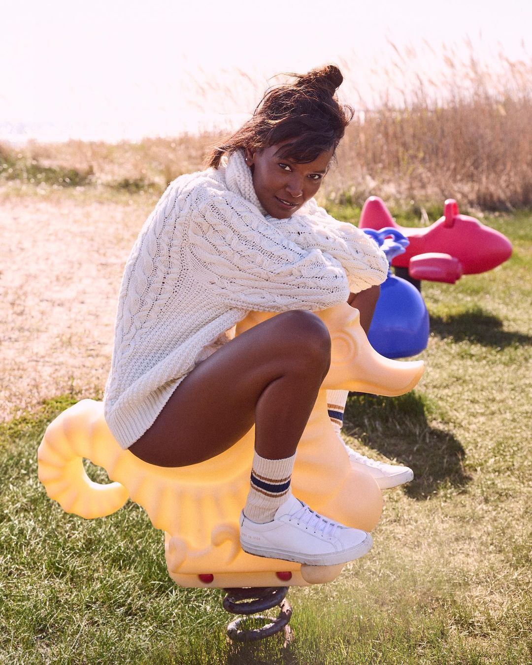 A woman with an updo sits on a yellow spring horse toy outside with a red airplane toy, wearing a white cable-knit sweater and sneakers.