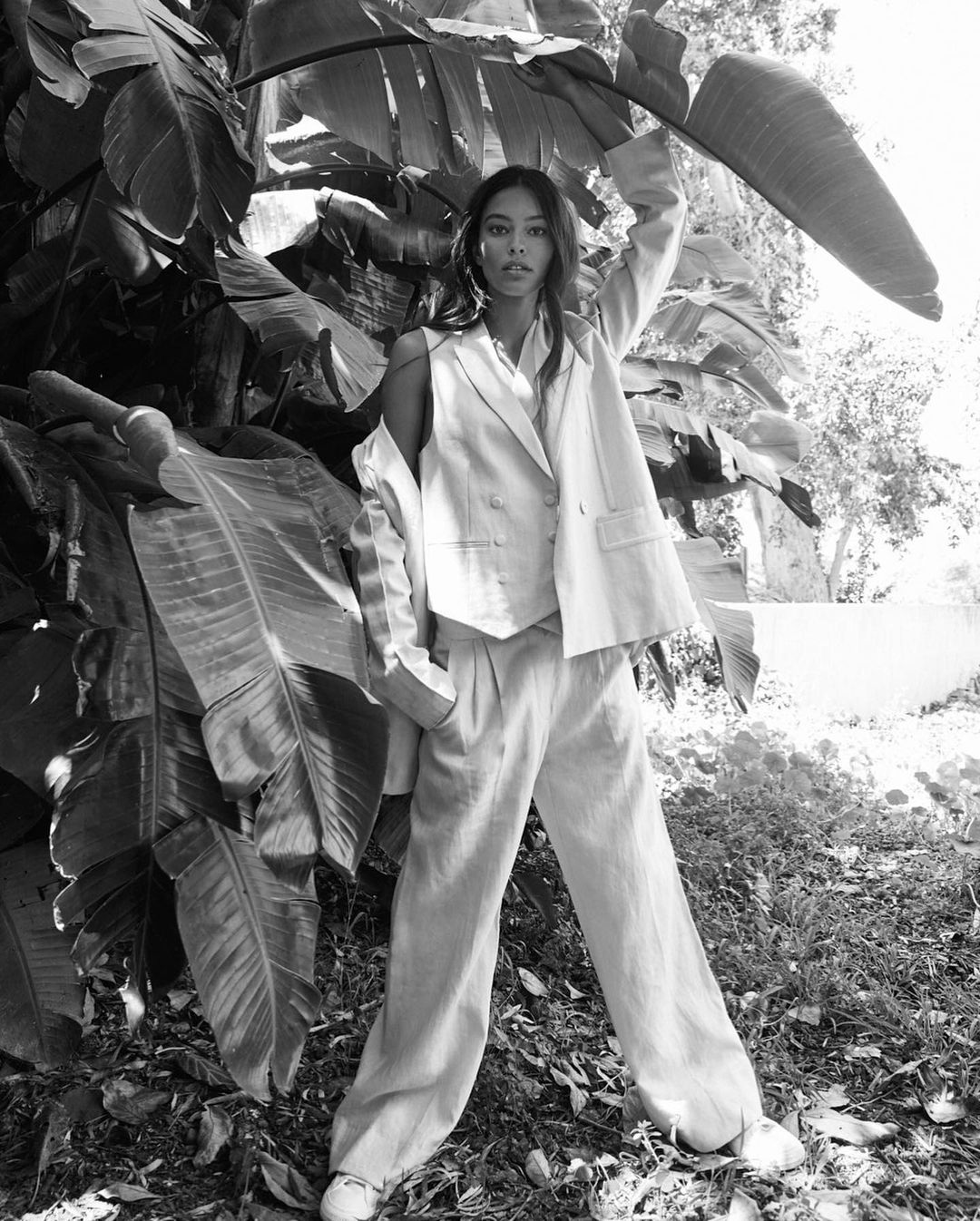 A woman in a suit poses near broadleaf plants outdoors in this full shot. The image, in black and white, features a high angle of light.