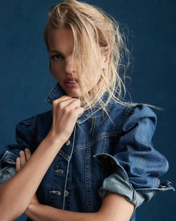 Portrait of a blonde woman wearing a blue denim jacket with puffed shoulders against a dark textured blue background.
