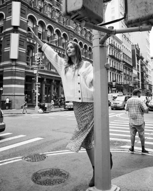 A woman in a cardigan and dress stands on a city street corner with buildings and traffic in the background in black and white.
