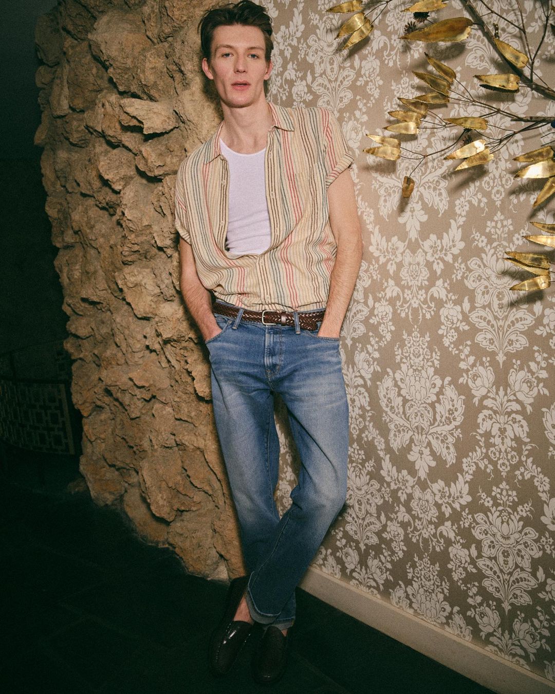Man in jeans and striped shirt leaning against patterned wall and rock texture, casual style, indoor setting, medium shot.